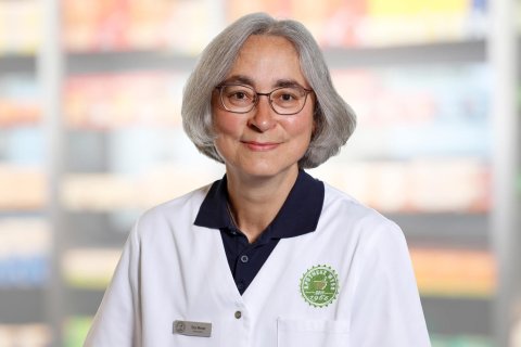 Ein Apotheker mit grauem Haar und Brille, der einen wei&szlig;en Laborkittel mit gr&uuml;nem Apothekenlogo tr&auml;gt, steht in einem Ladengang mit unscharfen Regalen mit Produkten im Hintergrund.