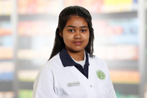 Eine junge Frau, Sumitha Sureshan, die einen wei&szlig;en Laborkittel und ein Namensschild tr&auml;gt, steht vor verschwommenen Regalen, m&ouml;glicherweise in einer Apotheke oder einem Gesch&auml;ft. Sie hat langes dunkles Haar und einen neutralen Gesichtsausdruck.