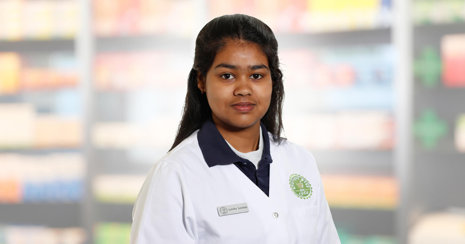 Eine junge Frau, Sumitha Sureshan, die einen wei&szlig;en Laborkittel und ein Namensschild tr&auml;gt, steht vor verschwommenen Regalen, m&ouml;glicherweise in einer Apotheke oder einem Gesch&auml;ft. Sie hat langes dunkles Haar und einen neutralen Gesichtsausdruck.