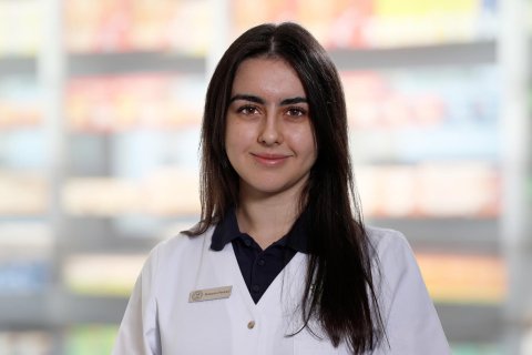 Eine junge Frau mit langen dunklen Haaren, die einen wei&szlig;en Laborkittel und ein Namensschild mit der Aufschrift Ekatarina Paclovic tr&auml;gt, steht l&auml;chelnd vor verschwommenen Regalen, die an eine Apotheke oder einen medizinischen Bereich erinnern.