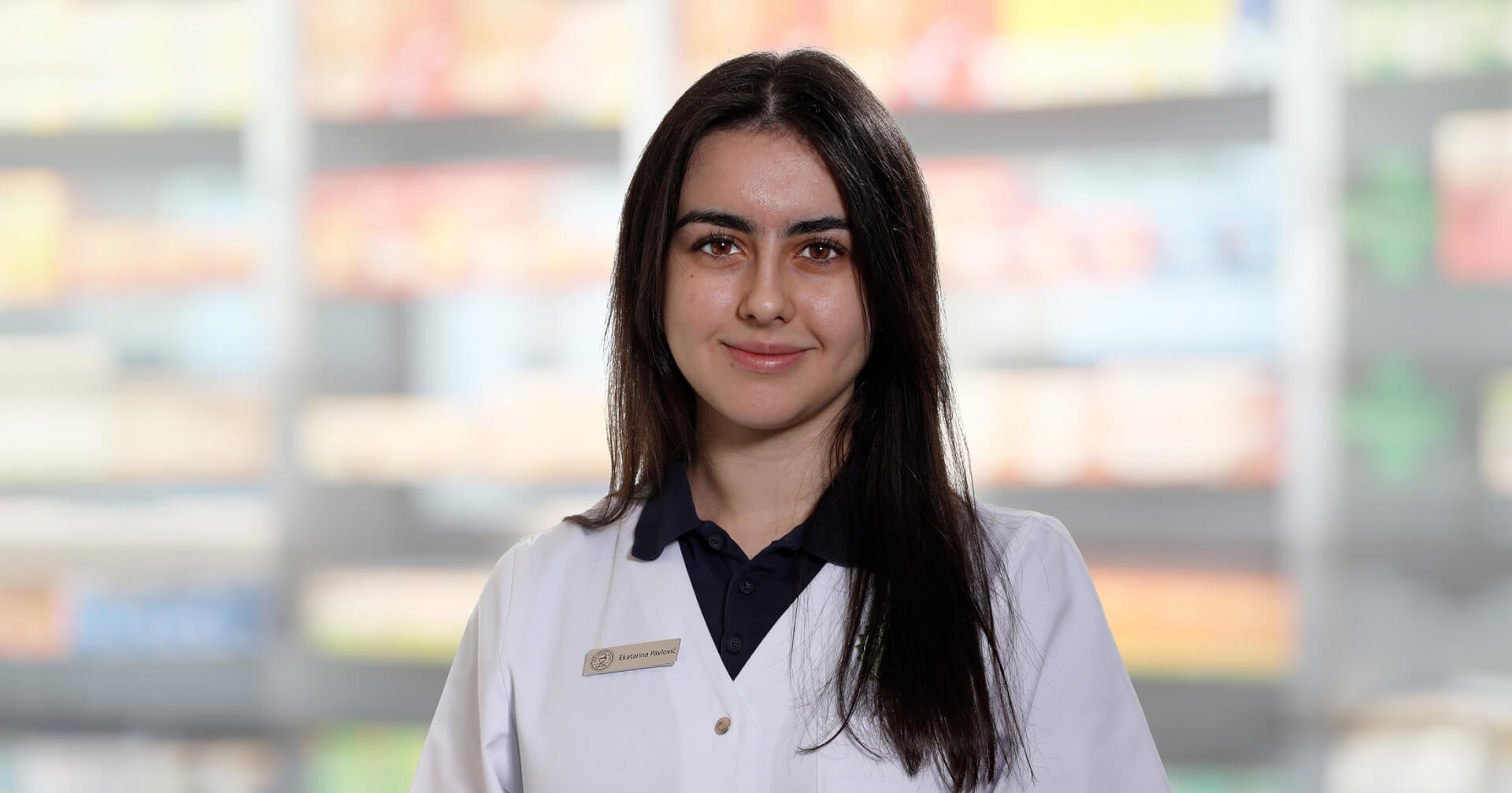 Eine junge Frau mit langen dunklen Haaren, die einen wei&szlig;en Laborkittel und ein Namensschild mit der Aufschrift Ekatarina Paclovic tr&auml;gt, steht l&auml;chelnd vor verschwommenen Regalen, die an eine Apotheke oder einen medizinischen Bereich erinnern.