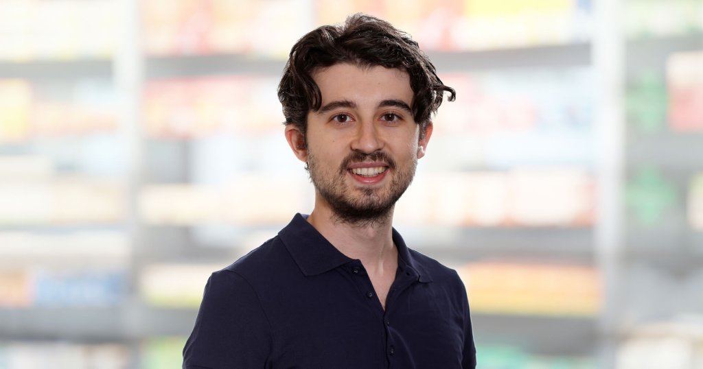 Ein junger Mann mit dunklem, gewelltem Haar und einem kurzen Bart l&auml;chelt in die Kamera und tr&auml;gt ein marineblaues Polohemd. Der Hintergrund ist unscharf und zeigt Regale mit bunten Produkten, die an einen Laden oder Supermarkt erinnern.
