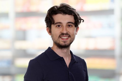 Ein junger Mann mit dunklem, gewelltem Haar und einem kurzen Bart l&auml;chelt in die Kamera und tr&auml;gt ein marineblaues Polohemd. Der Hintergrund ist unscharf und zeigt Regale mit bunten Produkten, die an einen Laden oder Supermarkt erinnern.