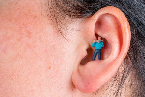 Eine Nahaufnahme eines menschlichen Ohrs mit einer digital hinzugef&uuml;gten winzigen Figur in einem blauen Hemd, die im Inneren des Geh&ouml;rgangs steht und ihn mit einem gro&szlig;en Wattest&auml;bchen zu reinigen scheint - als Symbol f&uuml;r den Wunsch, Nebenger&auml;usche aus dem t&auml;glichen Leben zu entfernen.