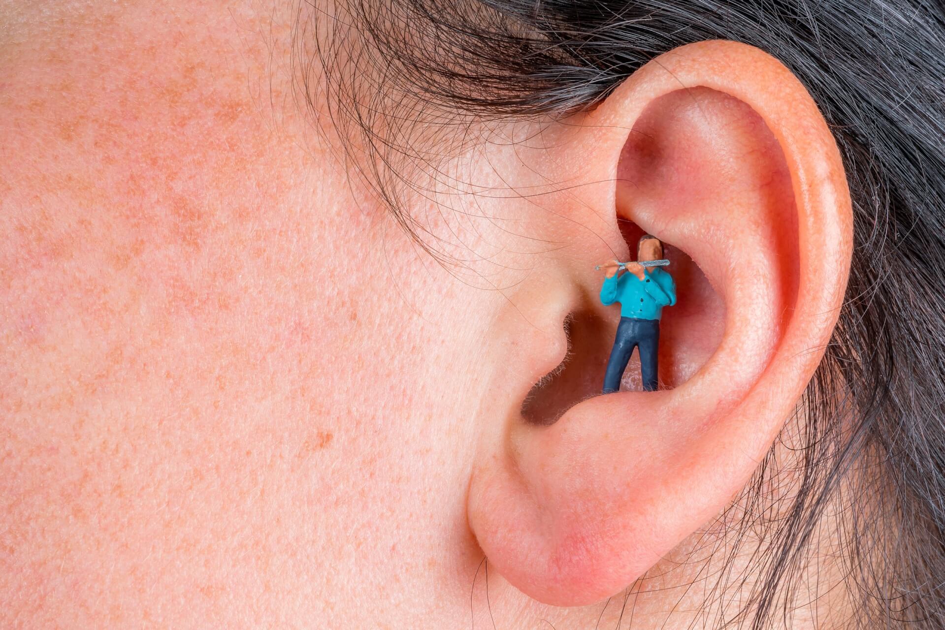 Eine Nahaufnahme eines menschlichen Ohrs mit einer digital hinzugef&uuml;gten winzigen Figur in einem blauen Hemd, die im Inneren des Geh&ouml;rgangs steht und ihn mit einem gro&szlig;en Wattest&auml;bchen zu reinigen scheint - als Symbol f&uuml;r den Wunsch, Nebenger&auml;usche aus dem t&auml;glichen Leben zu entfernen.