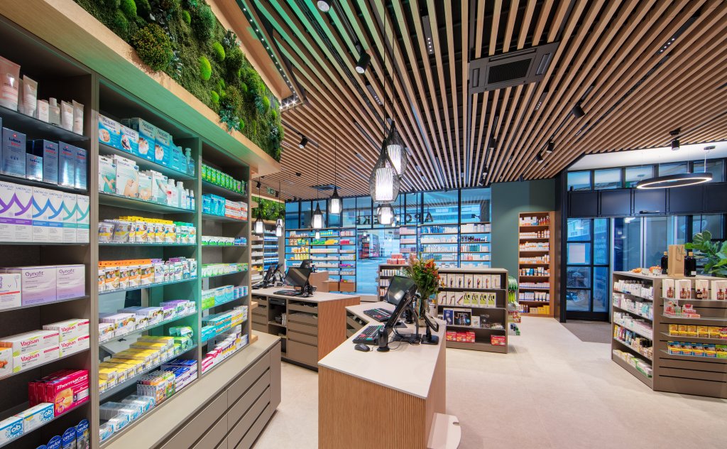 Modern gestaltete Apotheke Wyss nahe Bahnhof Baden mit Holzdeckenlamellen, beleuchteten Regalen voller Gesundheitsprodukte, einem geschwungenen Tresen mit Computern und gr&uuml;nen Pflanzen f&uuml;r ein frisches Ambiente.