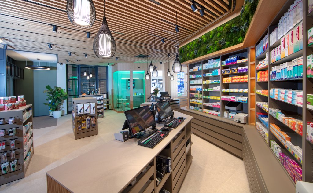 Modern gestaltet: Modernes Apothekeninterieur mit Holzdecke, Regalen mit Medikamenten und Gesundheitsprodukten in der Bahnhof Apotheke, zwei Computerregistern auf dem Tresen, einer gr&uuml;nen Pflanzenwand, heller Beleuchtung und einem im hinteren Bereich sichtbaren Beratungsbereich.
