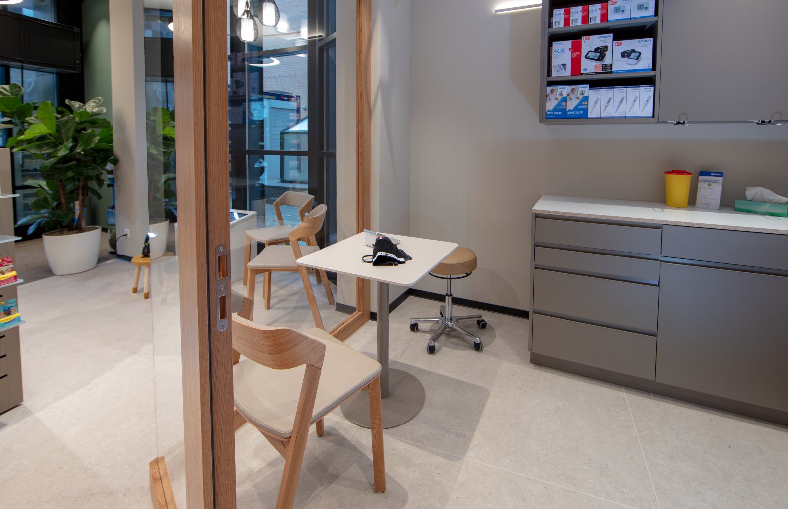 Ein modernes, sauberes Sprechzimmer in der Apotheke Wyss in der N&auml;he des Bahnhofs Baden ist mit Holzst&uuml;hlen, einem weissen Tisch, Schr&auml;nken und medizinischem Material ausgestattet. Gro&szlig;e Fenster und Zimmerpflanzen schaffen eine helle, freundliche Atmosph&auml;re.