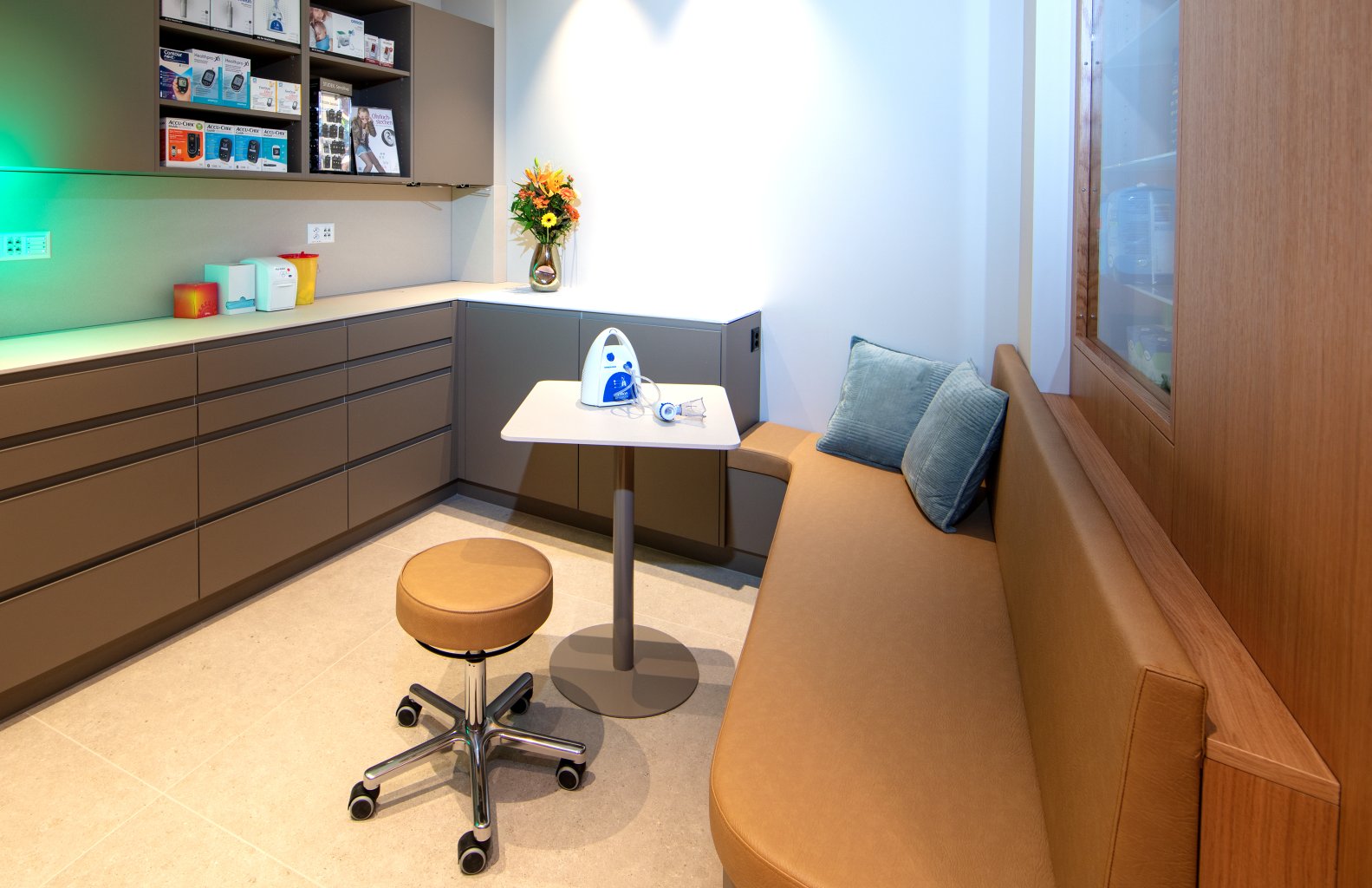 Eine moderne Arztpraxis in der N&auml;he des Bahnhofs Baden mit braunen Schr&auml;nken, einem Ecktresen mit medizinischem Material, einem kleinen Tisch mit einem Vernebler der Apotheke Wyss, einem Rollhocker, einer gepolsterten Sitzbank und einer Blumenvase bei heller Beleuchtung.
