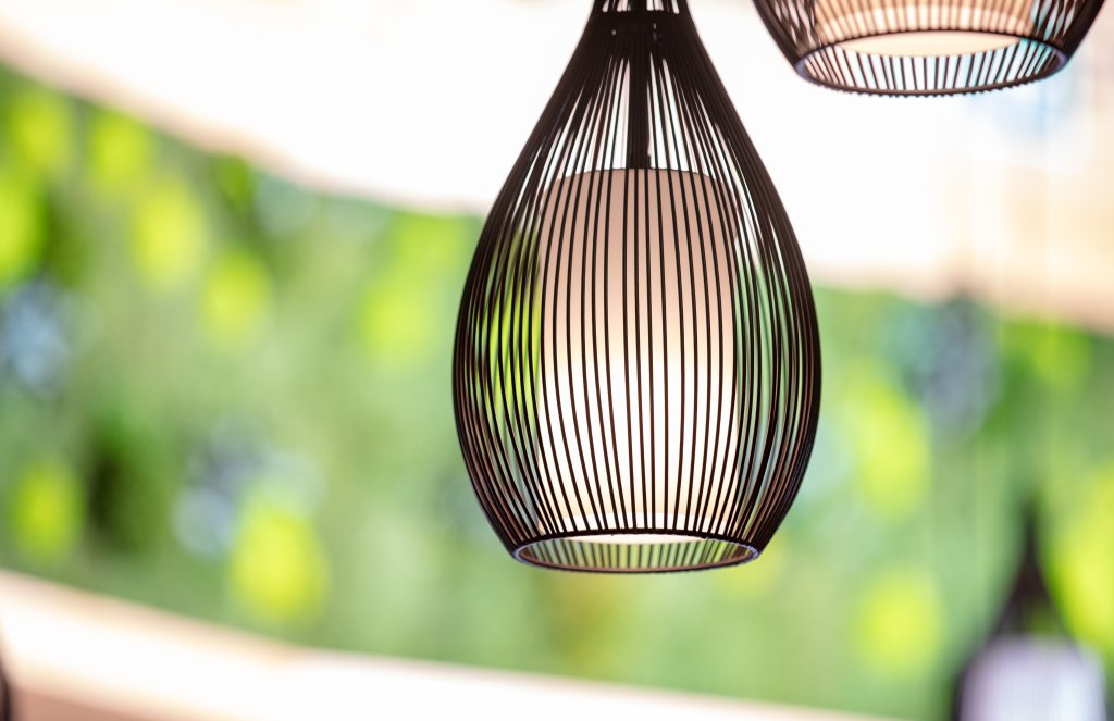 Nahaufnahme einer modernen Pendelleuchte mit schwarzem Drahtgitterdesign, die vor einem unscharfen gr&uuml;nen und gelben Hintergrund in der N&auml;he der Apotheke Wyss, Bahnhof Baden, h&auml;ngt.