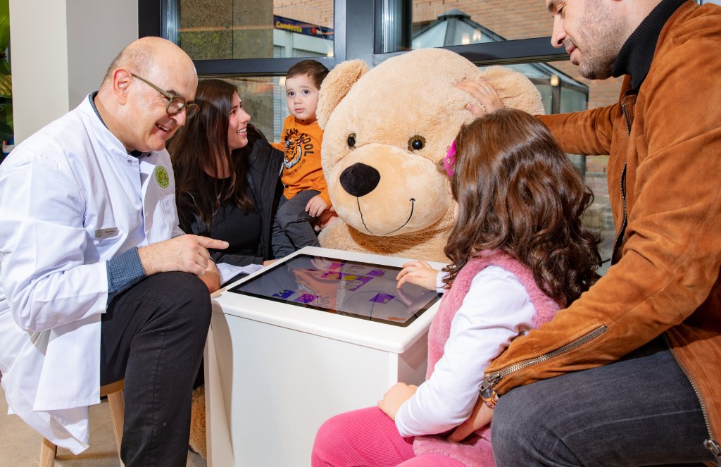 Ein Arzt, zwei Erwachsene und zwei Kinder sitzen in der frisch renovierten Apotheke Wyss in der N&auml;he des Bahnhofs Baden um einen Touchscreen-Tisch mit einem gro&szlig;en Teddyb&auml;ren in der Mitte; ein Kind interagiert mit dem Bildschirm, w&auml;hrend die Gruppe l&auml;chelt und sich miteinander besch&auml;ftigt.