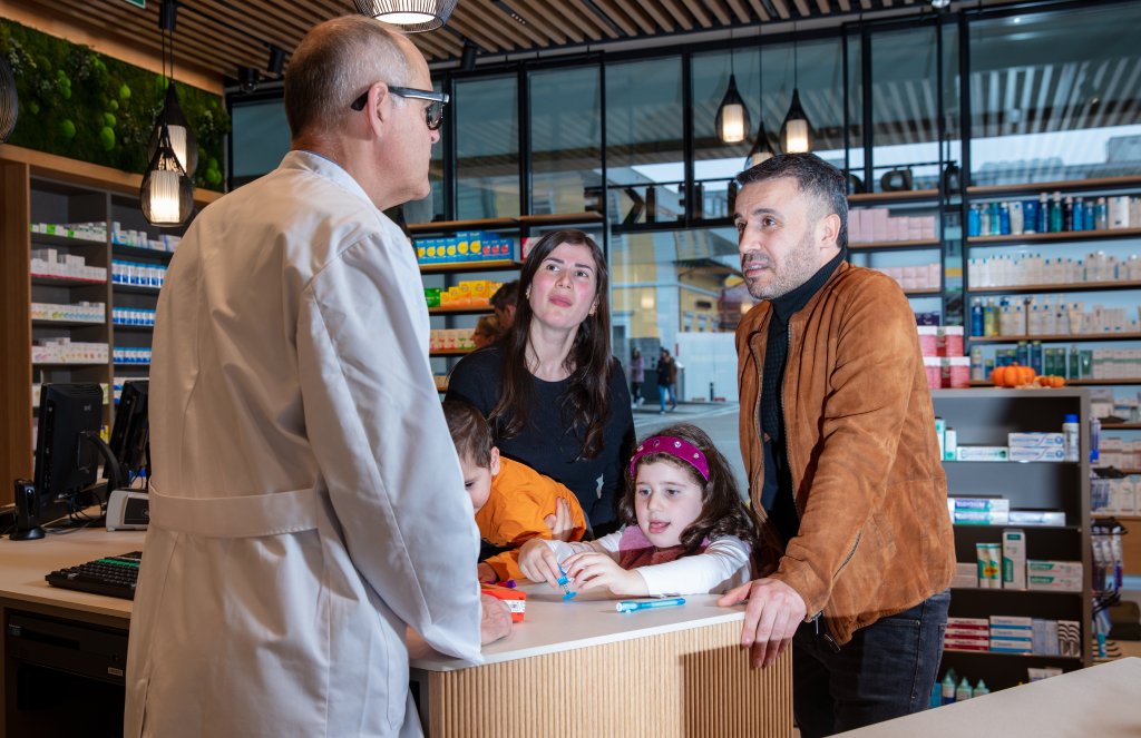 Eine Apothekerin im weissen Kittel spricht mit einem Mann, einer Frau und zwei Kindern in der modern gestalteten Apotheke Wyss am Bahnhof Baden. Die aufmerksame Familie steht an der Theke, im Hintergrund sind Regale mit Medikamenten zu sehen.