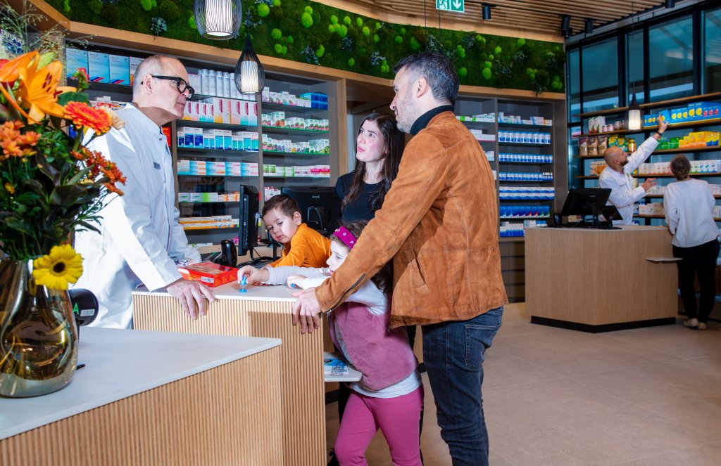 Eine Familie mit zwei Kindern spricht mit einer Apothekerin in der modern gestalteten Apotheke Wyss im Bahnhof Baden. Im Hintergrund stehen Regale mit Medikamenten und Gesundheitsprodukten und eine weitere Apothekerin bedient eine Kundin.