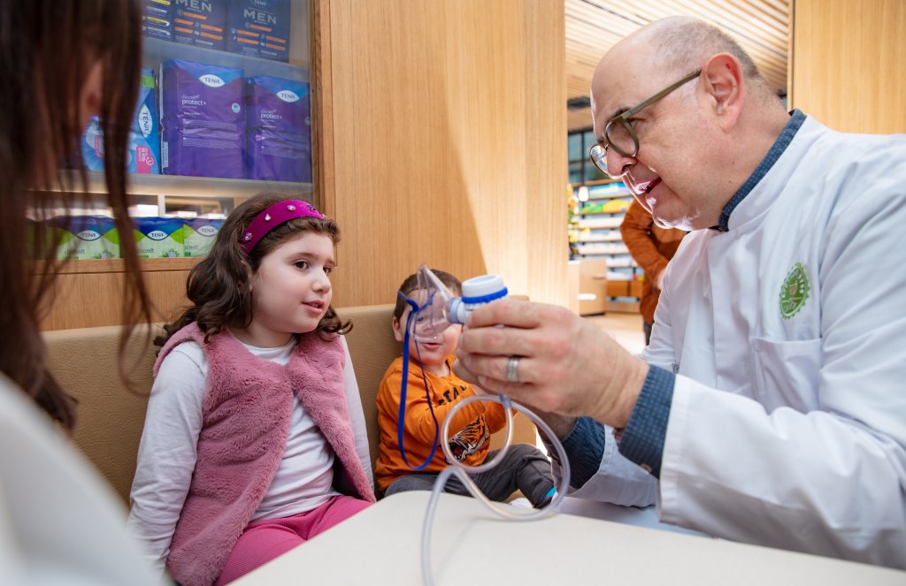Ein Apotheker im weissen Kittel in der Apotheke Wyss, Bahnhof Baden, zeigt einem M&auml;dchen und einem Jungen, die auf einer Bank sitzen, ein modernes medizinisches Ger&auml;t. Die Kinder schauen aufmerksam zu, w&auml;hrend links ein weiterer Erwachsener teilweise sichtbar ist.