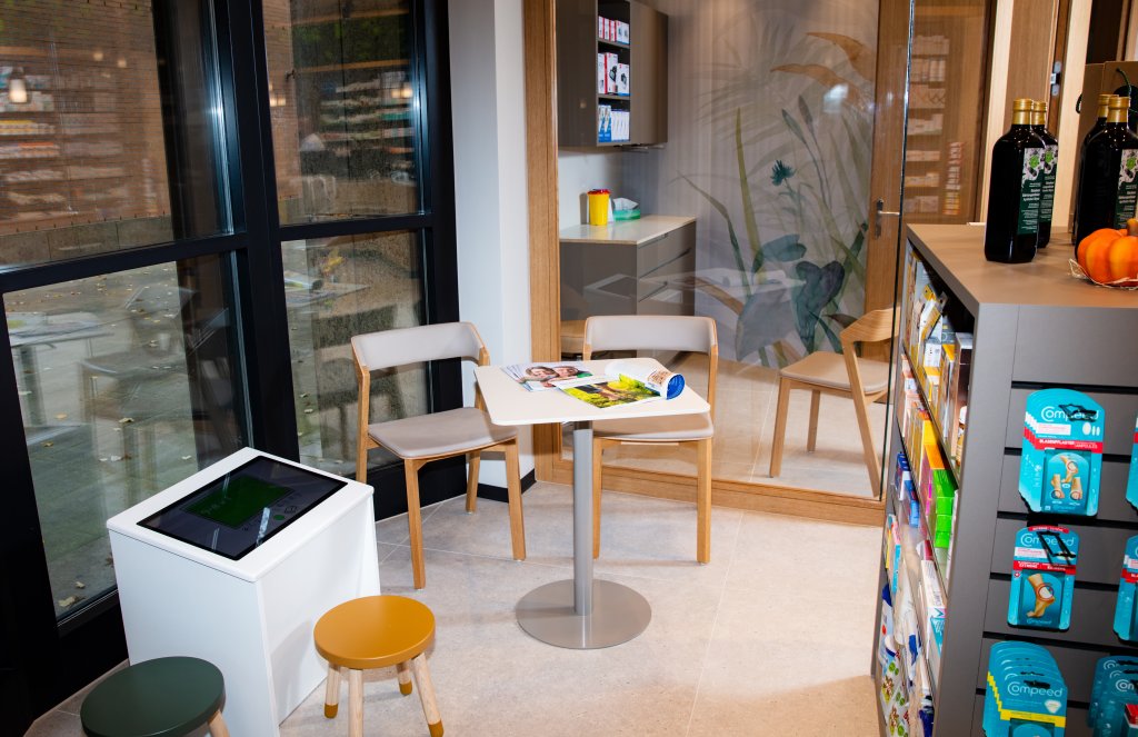 Ein kleiner, modern gestalteter Wartebereich in der Apotheke Wyss beim Bahnhof Baden ist mit einem Tisch, vier St&uuml;hlen, Zeitschriften, einem digitalen Touchscreen-Kiosk und einem Regal mit Gesundheitsprodukten ausgestattet. Gro&szlig;e Fenster und eine Glaswand mit Naturmotiven schaffen eine helle Atmosph&auml;re.