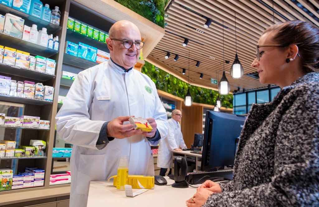 Ein Apotheker im weissen Kittel l&auml;chelt, als er in der modernen Apotheke Wyss am Bahnhof Baden einer Kundin &uuml;ber den Tresen hinweg eine Medikamentenschachtel zeigt, w&auml;hrend im Hintergrund hell erleuchtete Regale mit Produkten stehen.