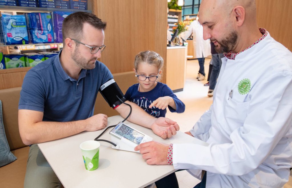 Ein Mann l&auml;sst sich in der Apotheke Wyss, Bahnhof Baden, von einer Fachperson den Blutdruck messen, w&auml;hrend ein junges M&auml;dchen in der hellen, modern gestalteten Umgebung neugierig auf den Monitor zeigt.