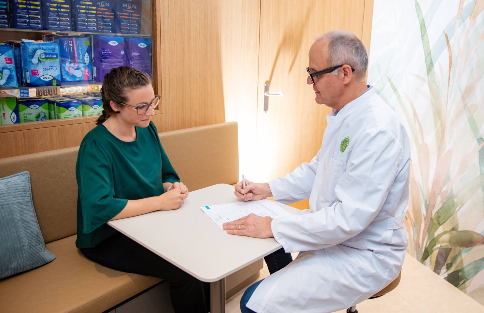 Ein Arzt im weissen Kittel sitzt an einem Tisch und schreibt in ein Formular, während er sich mit einer Frau mit Brille und grünem Hemd in der modern gestalteten Arztpraxis der Apotheke Wyss beim Bahnhof Baden unterhält. Im Hintergrund sind Regale mit Hygieneartikeln zu sehen.