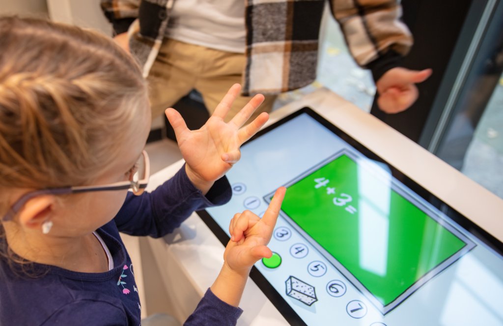 Ein junges M&auml;dchen z&auml;hlt mit den Fingern, w&auml;hrend es die Gleichung 4 + 3 = auf einem modernen Touchscreen-Ger&auml;t in der N&auml;he des Bahnhofs Baden l&ouml;st, w&auml;hrend ein anderes Kind daneben steht.