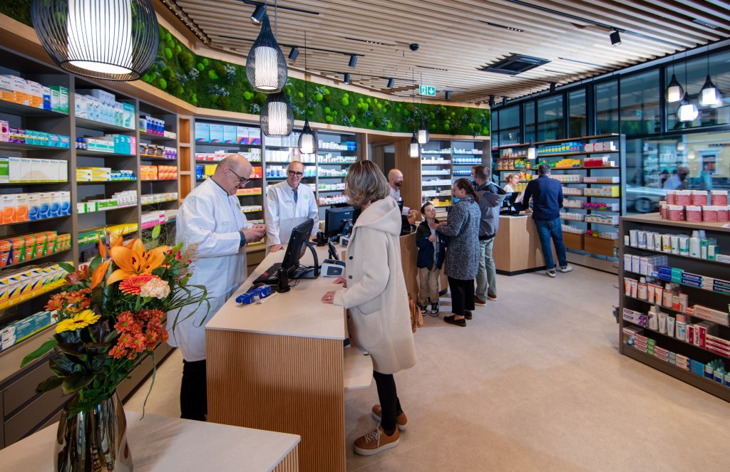 Eine modern gestaltete Apotheke Wyss am Bahnhof Baden, mit Pharmazeuten in wei&szlig;en Kitteln, die Kunden am Tresen beraten. Regale voller Medikamente s&auml;umen die W&auml;nde, w&auml;hrend Blumen den Tresen schm&uuml;cken und Kunden in der Schlange warten.