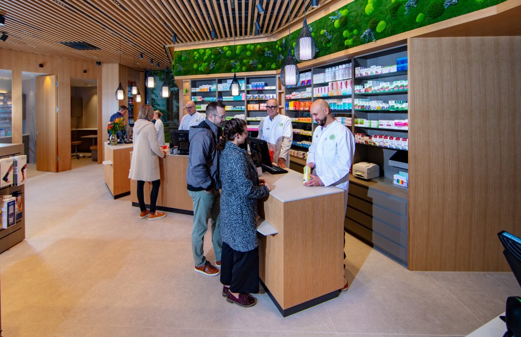 In der modernen Apotheke Wyss am Bahnhof Baden stehen die Menschen an den Schaltern und sprechen mit den Apothekerinnen und Apothekern. An den W&auml;nden stehen Regale mit Medikamenten, der Raum ist mit Holzakzenten und gr&uuml;nen Pflanzen geschm&uuml;ckt.