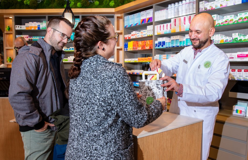 In der Apotheke Wyss &uuml;berreicht eine Apothekerin einer l&auml;chelnden Frau am modernen Apothekentresen in der N&auml;he des Bahnhofs Baden eine T&uuml;te mit Medikamenten. Im Hintergrund stehen bunte Medikamentenschachteln in den Regalen.