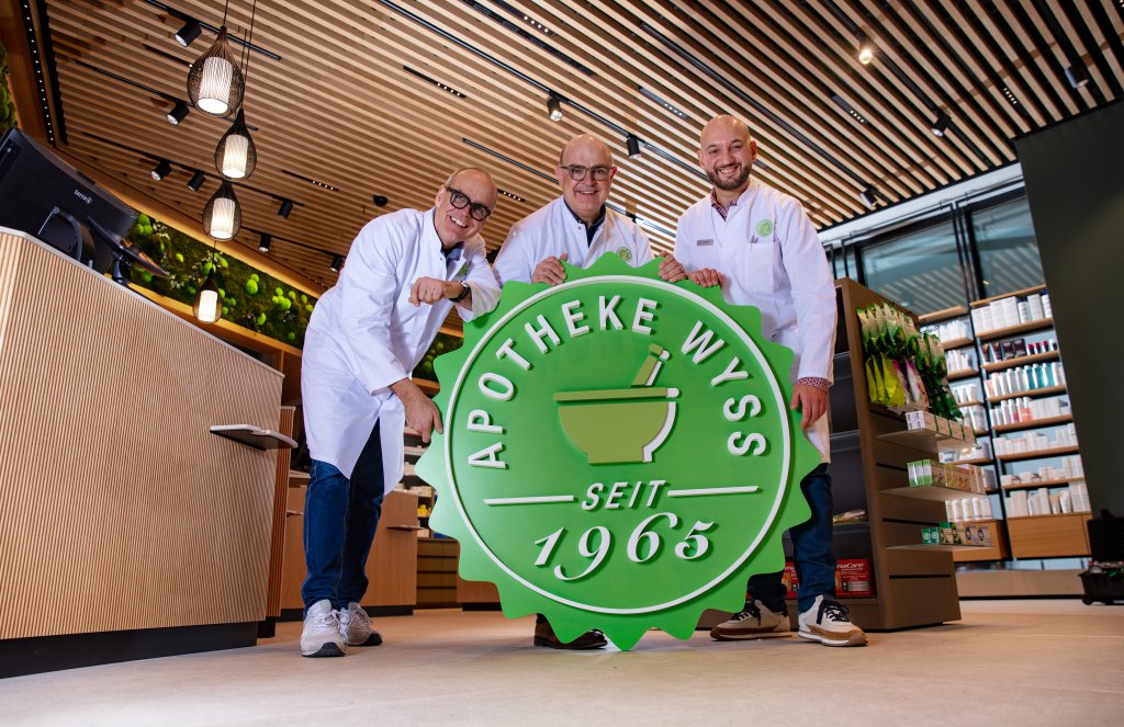 Drei l&auml;chelnde Personen in wei&szlig;en Laborkitteln halten ein gro&szlig;es gr&uuml;nes Schild APOTHEKE WYSS SEIT 1965 im Inneren einer modernen Apotheke am Bahnhof Baden, im Hintergrund sind Regale mit Produkten zu sehen.