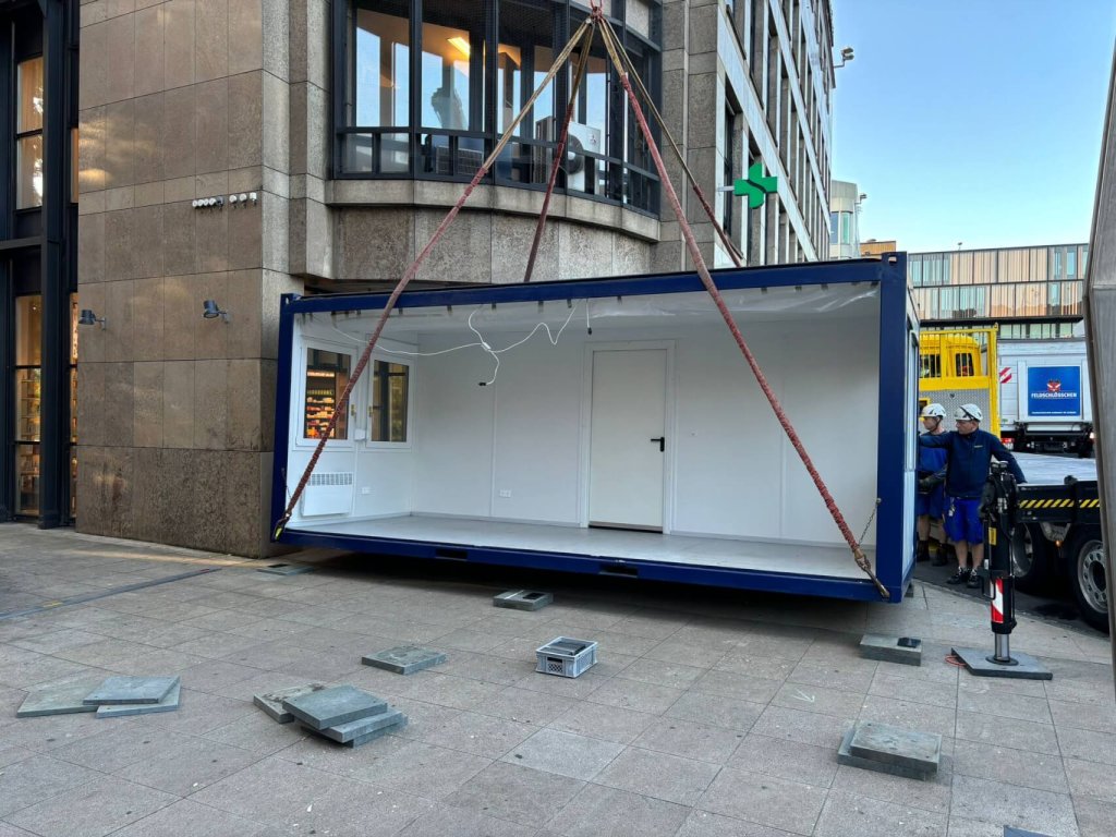 Vor dem modern gestalteten Bahnhof Baden wird ein grosser, offener Schiffscontainer an Seilen hochgezogen. Mehrere Arbeiter in Schutzausr&uuml;stung stehen in der N&auml;he, Baumaterialien liegen auf dem Boden bei der Apotheke Wyss verstreut.