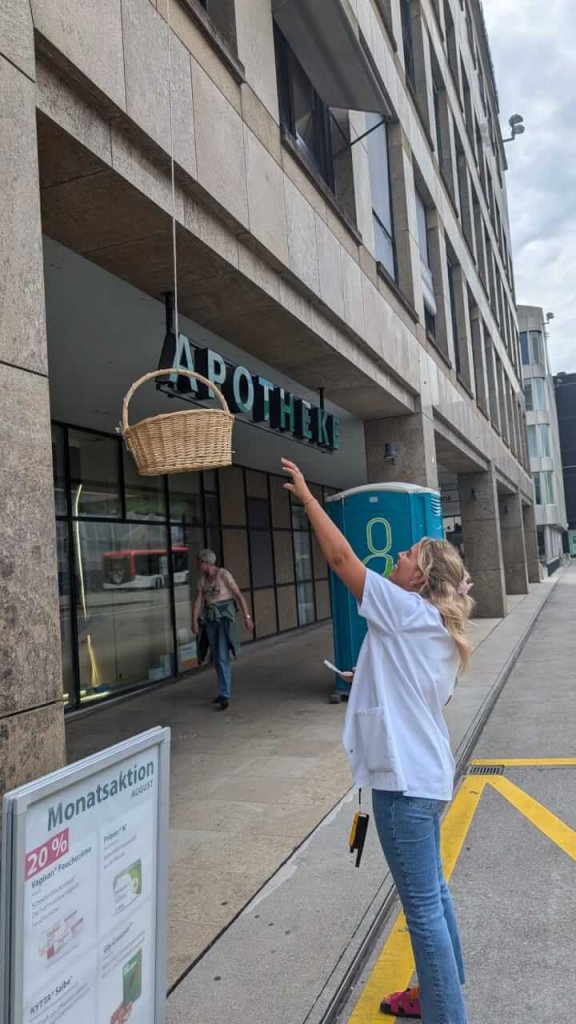 Eine Frau steht vor der modernen Apotheke Wyss in Baden und greift nach einem Korb, der an einem Seil herabgelassen wird. Eine andere Person geht im Hintergrund neben einer blauen mobilen Toilette vorbei.