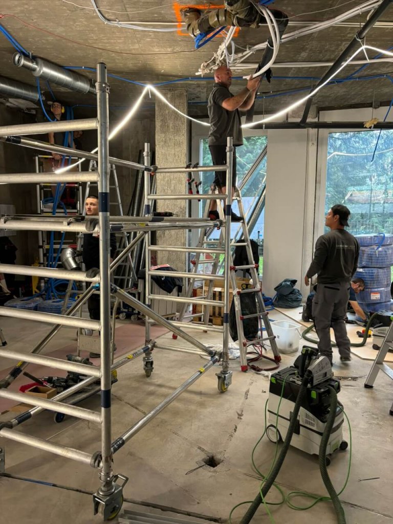 Vier Arbeiter installieren oder reparieren elektrische Leitungen und Beleuchtung an der Decke eines unfertigen Raums im Bahnhof Baden. Zwei von ihnen benutzen Leitern und Ger&uuml;ste, w&auml;hrend zwei andere in der N&auml;he arbeiten. Werkzeuge und Baumaterialien liegen auf der Baustelle verstreut.