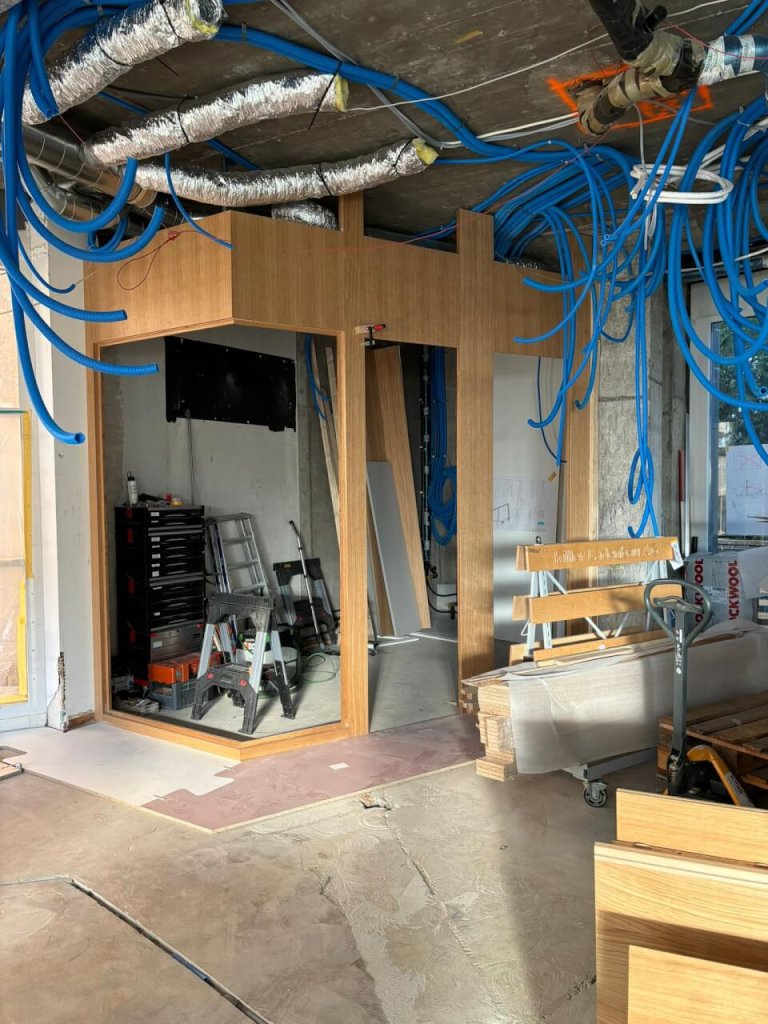 Ein moderner Baubereich mit freiliegenden Rohren und blauen Kabeln, die von der Decke h&auml;ngen. Eine Holzkonstruktion mit Glasscheiben, Werkzeug, eine Leiter und Baumaterialien liegen im unfertigen Raum des Bahnhofs Baden verstreut.