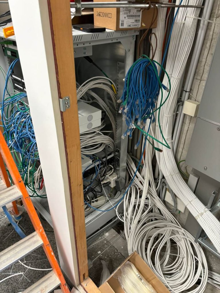 Ein Netzwerkschrank, aus dem blaue und weisse Kabel herausquellen, umgeben von weiteren losen Kabeln auf dem Boden und in Regalen, neben einer Leiter und einer Wand mit Steckdosen in der modern gestalteten Apotheke Wyss Baden.