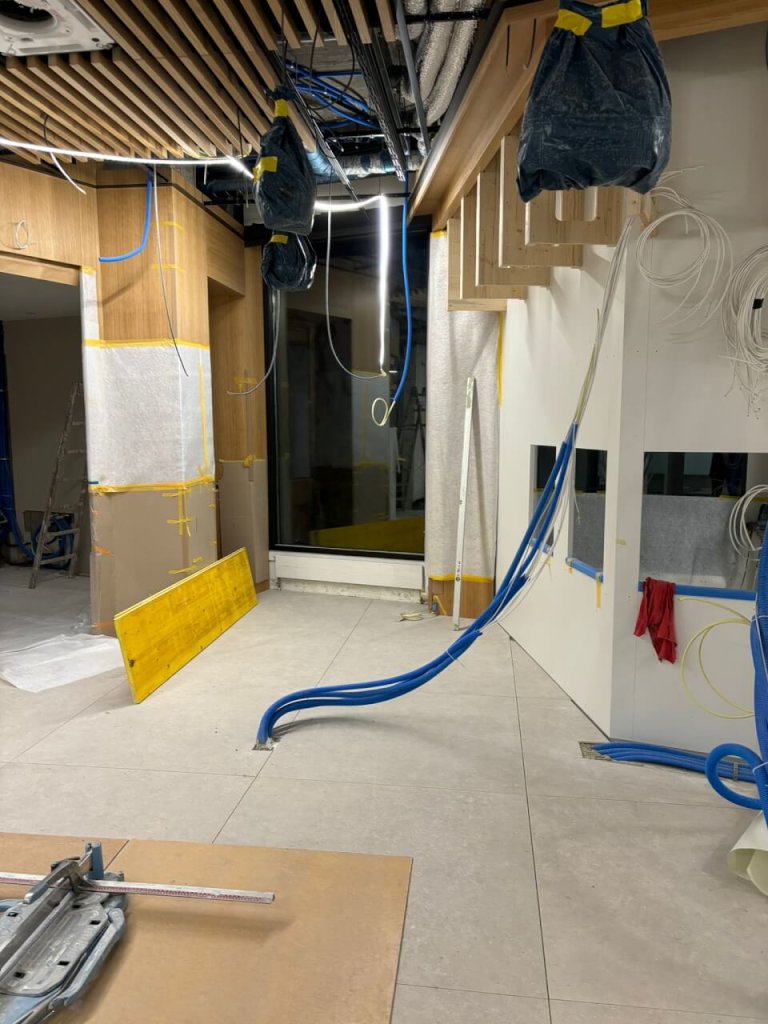 Ein im Bau befindlicher Innenraum der Apotheke Wyss, Bahnhof Baden, ist modern gestaltet mit freiliegenden Dr&auml;hten, blauen Kabeln, die von der Decke und den W&auml;nden h&auml;ngen, abgedeckten Armaturen und verstreuten Werkzeugen inmitten von abgeklebten Schutzbereichen.