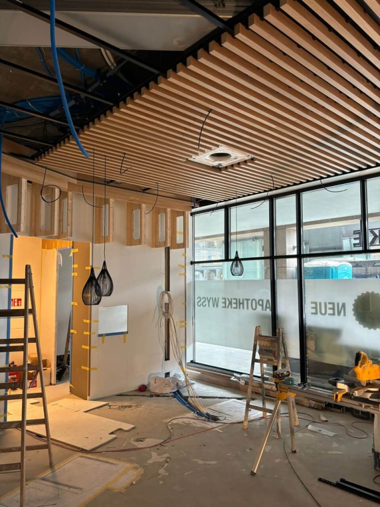 Ein modern gestalteter Innenraum von Apotheke Wyss am Bahnhof Baden im Bau, mit Holzlamellendecke, offenen Kabeln, Leitern, Werkzeugen und unfertigen W&auml;nden. Gro&szlig;e Fenster lassen Beschilderung und Tageslicht herein.