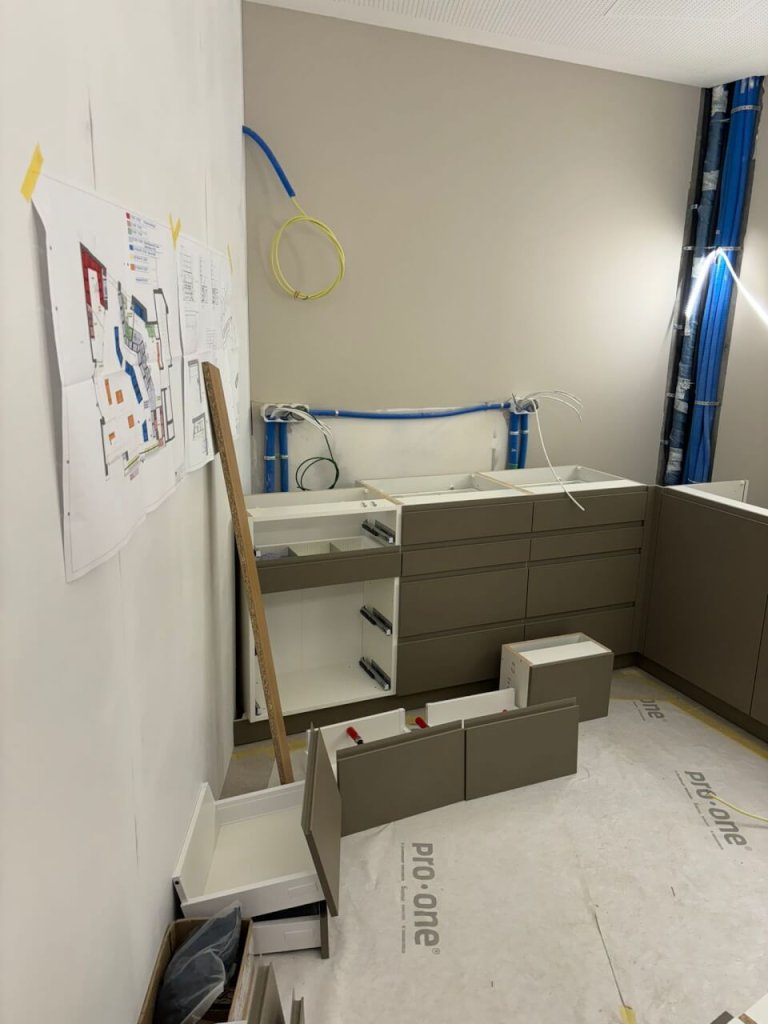 Eine moderne, teilweise installierte K&uuml;che mit hellbraunen Schr&auml;nken, offenen Schubladen und losen Schubladen auf dem Boden. Im Bahnhof Baden h&auml;ngen Baupl&auml;ne an der Wand, &uuml;ber den Schr&auml;nken sind Kabel und Rohre zu sehen.