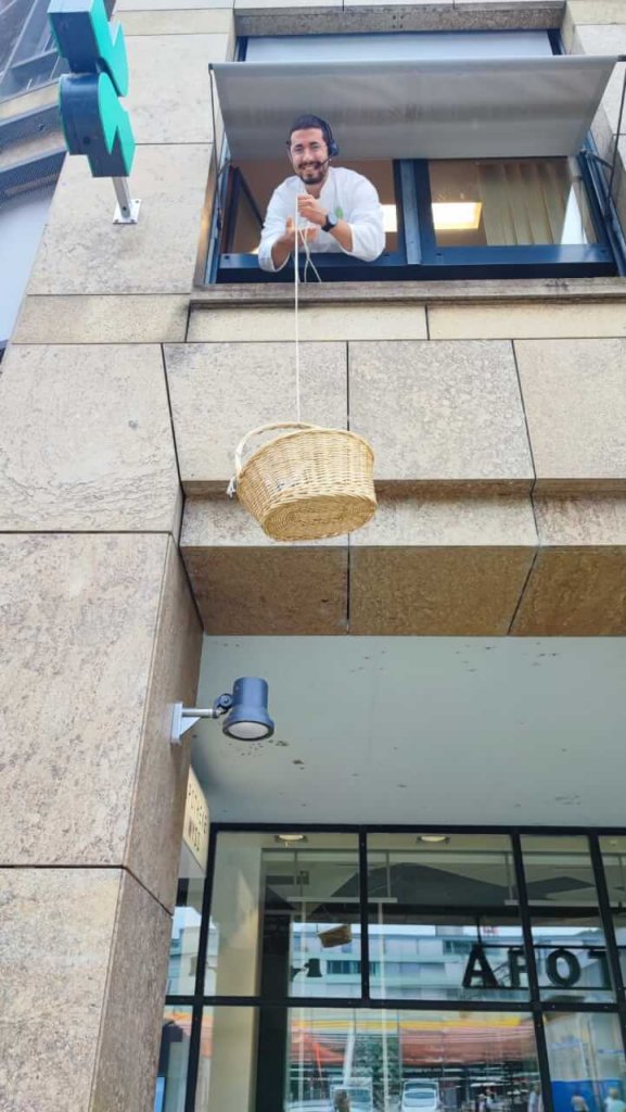 Ein l&auml;chelnder Mann mit einem Headset lehnt sich aus einem Fenster im oberen Stockwerk eines frisch renovierten modernen Geb&auml;udes und l&auml;sst einen Weidenkorb an einer Schnur in der N&auml;he des Bahnhofs Baden zu Boden.