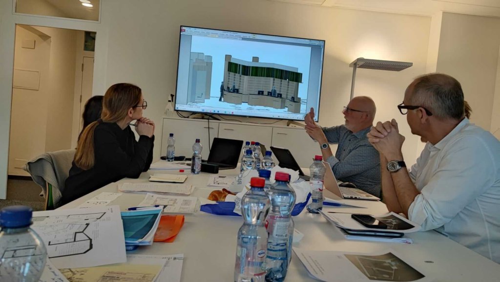 Drei Personen sitzen um einen mit Papieren und Wasserflaschen bedeckten Tisch und diskutieren &uuml;ber ein modern gestaltetes Geb&auml;udemodell f&uuml;r die Apotheke Wyss beim Bahnhof Baden, das auf einem grossen Bildschirm gezeigt wird. Eine Person gestikuliert in Richtung des Bildschirms, w&auml;hrend die anderen aufmerksam zuh&ouml;ren.