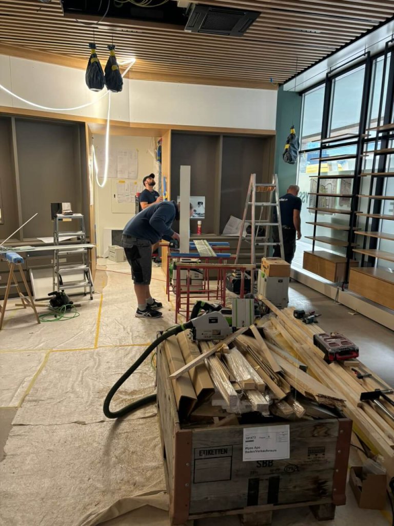 In einem teilweise errichteten, modern gestalteten Raum f&uuml;r die neue Apotheke Wyss in der N&auml;he des Bahnhofs Baden sind mehrere Arbeiter dabei, Ger&auml;te einzurichten und mit Werkzeugen zu arbeiten.
