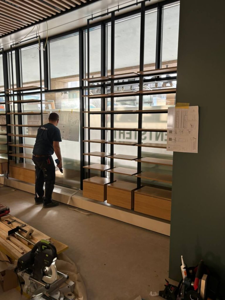 Eine Person arbeitet in einem modern gestalteten Raum in der N&auml;he gro&szlig;er Fenster und montiert oder &uuml;berpr&uuml;ft Holzregale, die an einer gr&uuml;nen Wand befestigt sind. Werkzeuge und Baumaterialien liegen verstreut auf dem Boden. In der N&auml;he sind Pl&auml;ne an die Wand geklebt.