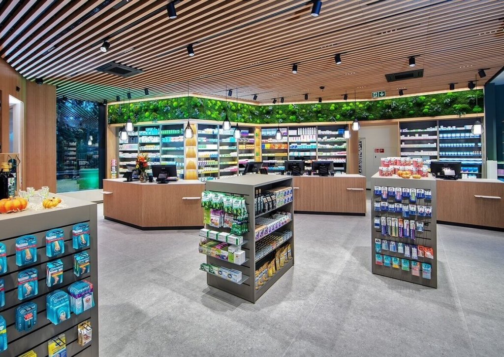 Moderne Apothekeneinrichtung mit h&ouml;lzernen Akzenten, gr&uuml;nem Wanddekor und gut organisierten Regalen mit Medikamenten, Gesundheitsprodukten und Toilettenartikeln. Mehrere Theken und helle Beleuchtung schaffen eine saubere, einladende Atmosph&auml;re am Standort Baden.