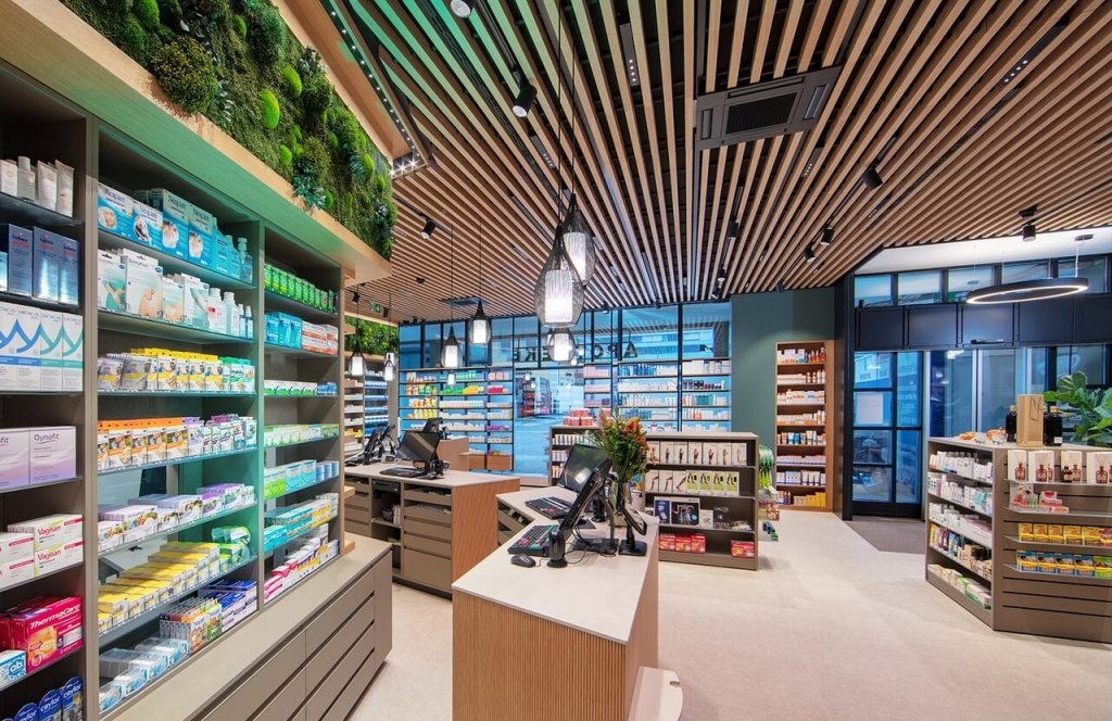 Modernes Apothekeninterieur am Standort Baden mit h&ouml;lzernen Deckenleisten, Regalen mit Medikamenten und Gesundheitsprodukten, digitalen Kassen am Tresen und Gr&uuml;npflanzen, die dem hellen, organisierten Raum einen nat&uuml;rlichen Touch verleihen.