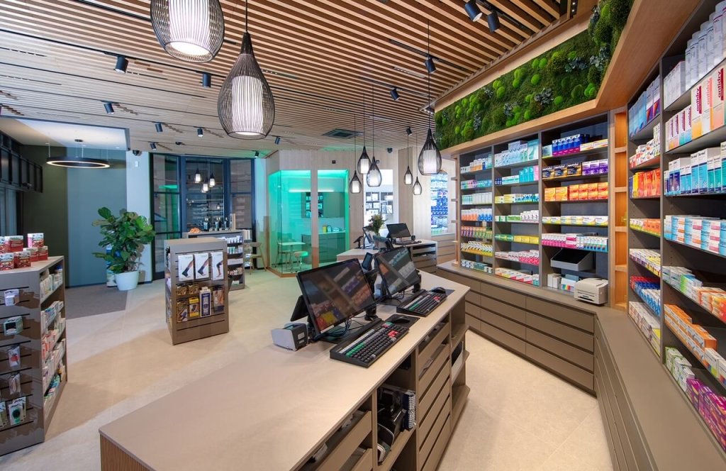 Moderne Apothekeneinrichtung am Standort Baden mit Holzdecke, Oberlichtern und Regalen mit bunten Medikamentenboxen. Auf dem Tresen stehen zwei Computerterminals, im Hintergrund ist ein gr&uuml;n beleuchteter Beratungsraum zu sehen.