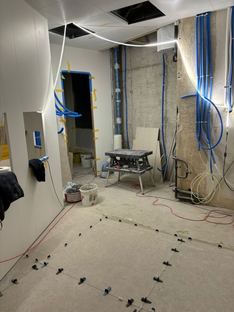 Ein im Bau befindlicher Raum in der Apotheke Wyss beim Bahnhof Baden, mit freiliegenden Rohren, unfertigen W&auml;nden, verstreuten Werkzeugen, sichtbarem Klebeband und Abstandshaltern, sowie einigen Fliesen, die gerade verlegt werden - der Raum wird bald modern gestaltet sein.