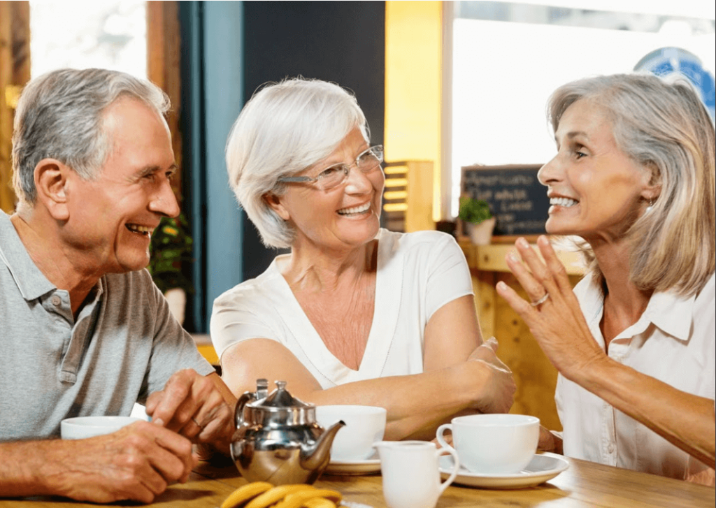 Drei &auml;ltere Erwachsene mit grauen Haaren sitzen an einem gem&uuml;tlichen Caf&eacute;tisch, l&auml;cheln und plaudern bei Tee und Bananen, wobei sie die Bedeutung sozialer Kontakte f&uuml;r die mentale Gesundheit reflektieren, w&auml;hrend sie die Gesellschaft des anderen genie&szlig;en.