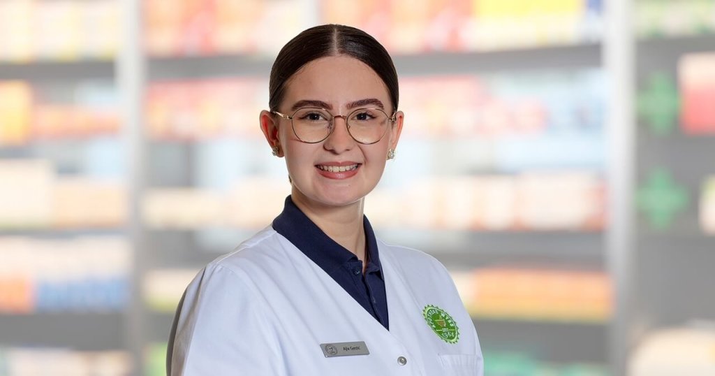 Eine junge Frau mit Brille und wei&szlig;em Laborkittel steht l&auml;chelnd vor den verschwommenen Apothekenregalen. Sie hat ihr dunkles Haar zur&uuml;ckgesteckt und tr&auml;gt ein Namensschild und einen gr&uuml;nen Apothekenausweis.