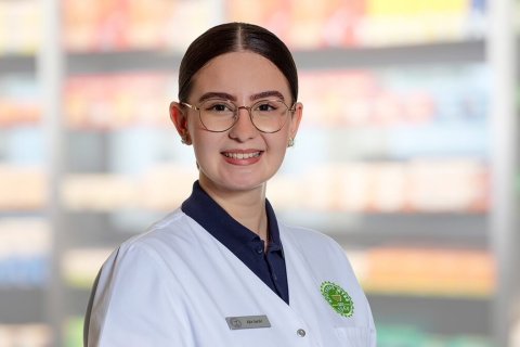 Eine junge Frau mit Brille und wei&szlig;em Laborkittel steht l&auml;chelnd vor den verschwommenen Apothekenregalen. Sie hat ihr dunkles Haar zur&uuml;ckgesteckt und tr&auml;gt ein Namensschild und einen gr&uuml;nen Apothekenausweis.