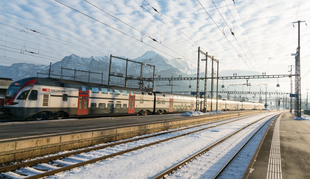 Ein moderner, auf Fahrt&uuml;chtigkeit gepr&uuml;fter Doppelstockzug steht an einem verschneiten Bahnsteig mit Bergen und blauem Himmel im Hintergrund; mehrere Gleise und Oberleitungen sind zu sehen.