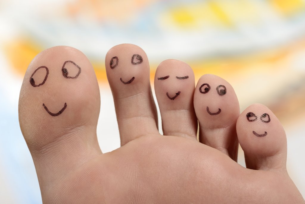 Eine Nahaufnahme von f&uuml;nf F&uuml;ssen mit fr&ouml;hlichen Gesichtern, die mit schwarzer Tinte gezeichnet sind, wobei jeder Zeh einen anderen Ausdruck zeigt. Der Hintergrund ist mit weichen Farben verschwommen, was den Eindruck erweckt, dass diese fr&ouml;hlichen Zehen in guten H&auml;nden sind.