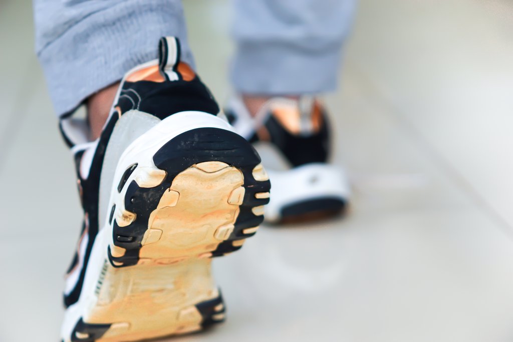 Nahaufnahme von F&uuml;ssen in Turnschuhen, w&auml;hrend eine Person auf einem gl&auml;nzenden, hellen Boden geht. Hellgraue Hosen vervollst&auml;ndigen die Szene und betonen die Bewegung und das Schuhwerk in guten H&auml;nden.