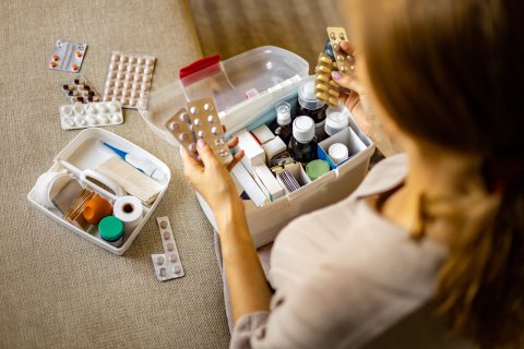 Eine Person sitzt auf einer Couch und sortiert Tabletten und medizinisches Material, h&auml;lt Blisterpackungen mit Tabletten in der Hand, w&auml;hrend zwei ge&ouml;ffnete Erste-Hilfe-K&auml;sten und verschiedene Medikamente auf dem Tisch ausgebreitet sind - damit ihre Hausapotheke f&uuml;r ein gesundes und fittes Leben bereit ist.